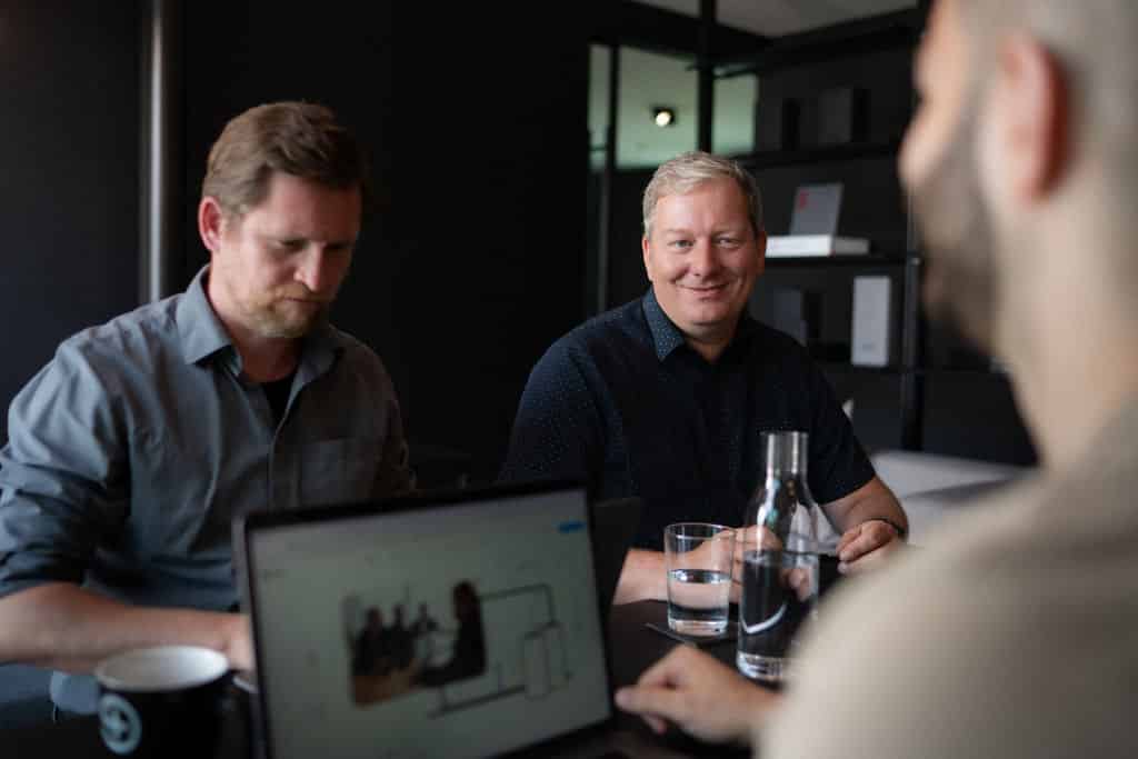 Troi Meetingfoto: Drei Personen am Konferenztisch mit Laptop und Getränken, im Gespräch und Austausch
